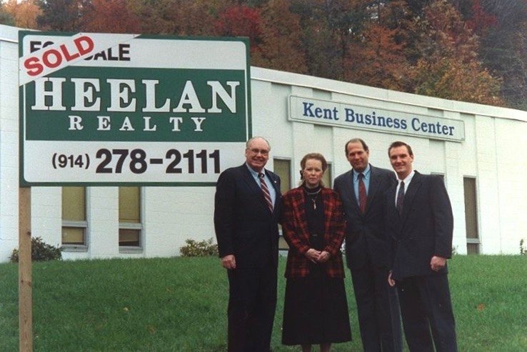 Archival photograph of four people standing beside a Heelan Realty Sold sign in front of a Hudson Valley commercial building.